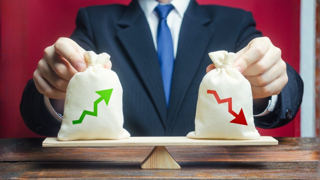 A businessman in a suit balancing two money bags with green upward and red downward profit arrows on a wooden scale to represent financial risk management.