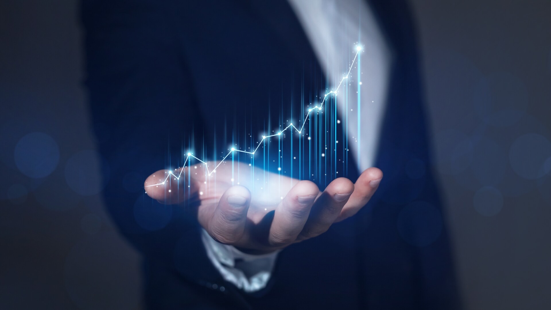 A close-up of a businessman in a dark suit holding a glowing blue digital line chart showing upward financial growth and market trends.