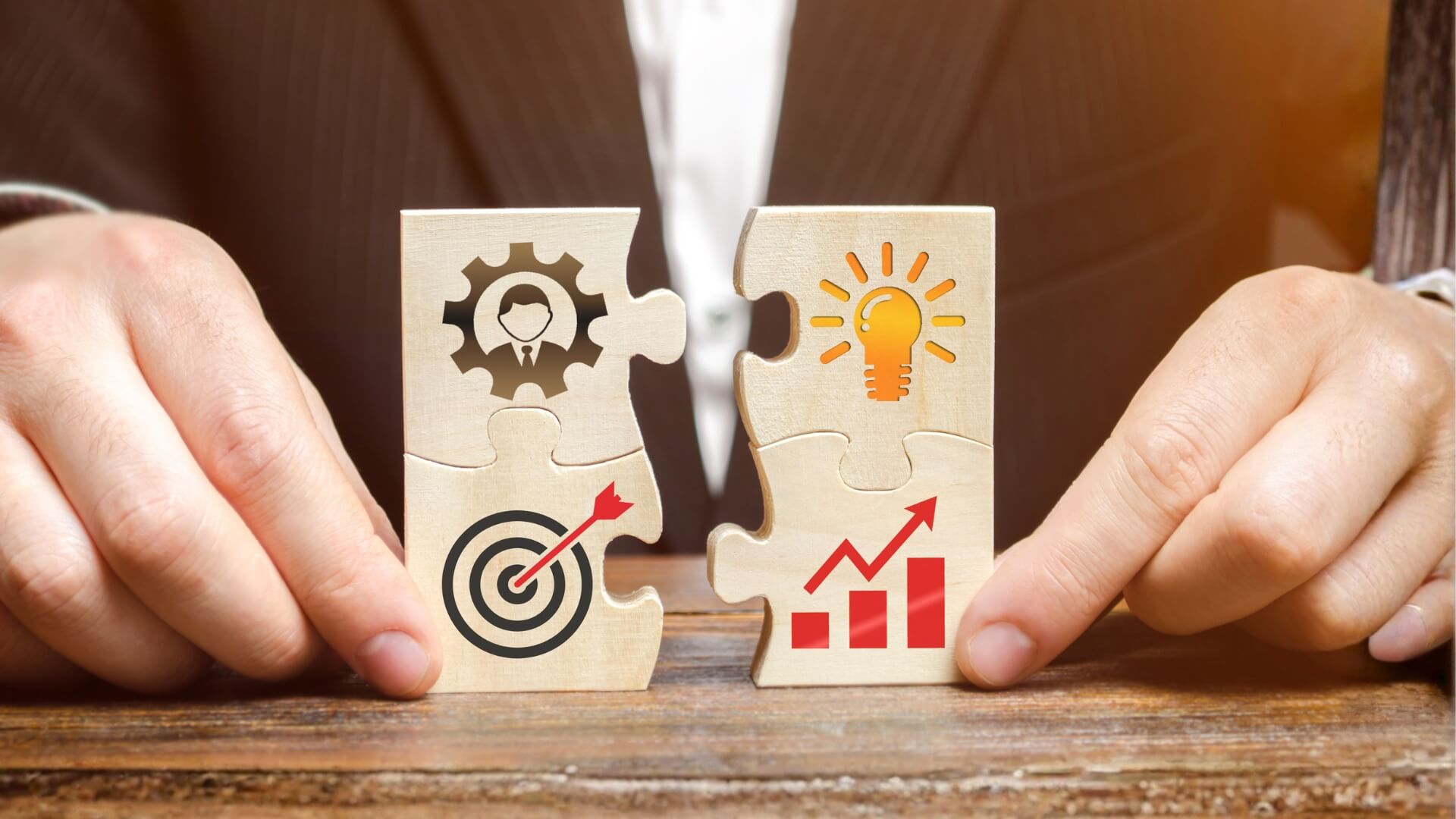A businessman's hands joining two wooden puzzle pieces featuring icons for corporate management, marketing target, innovative idea lightbulb, and financial growth chart.