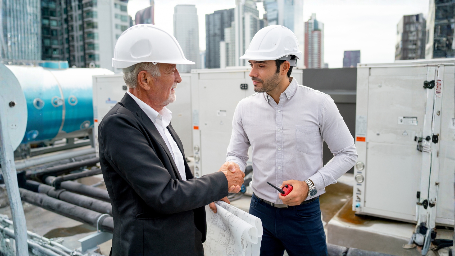 A building investor and an HVAC engineer in white hard hats shaking hands during an inspection of industrial rooftop air conditioning units.